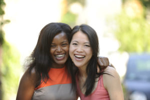 Two girls laughing with arms over each other's shoulders. On the left a woman of colour and on the right an Asian woman.  